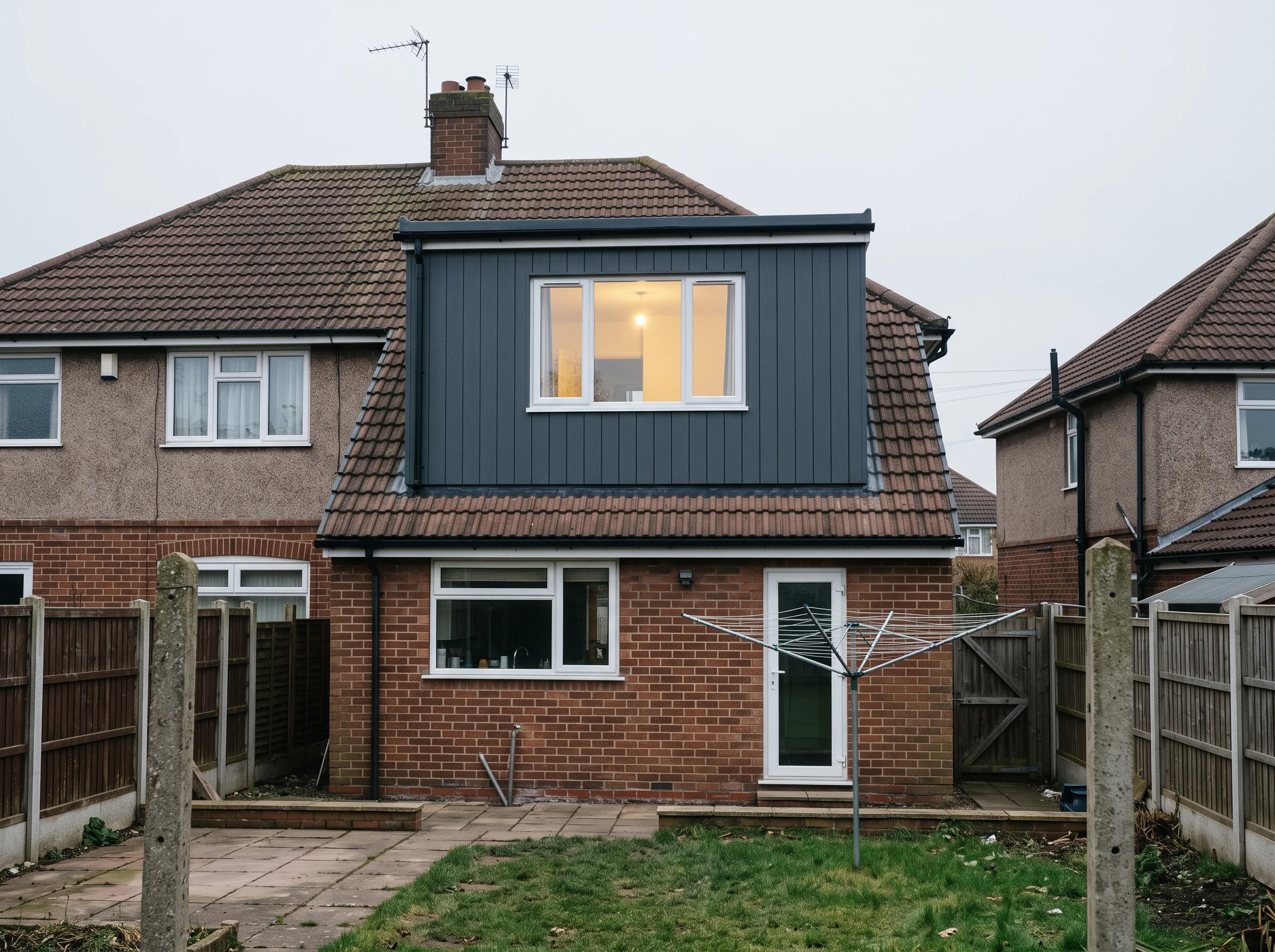 A brick semi-detached house with a dark grey dormer loft conversion and a lit window, viewed from the back garden with a rotary washing line.